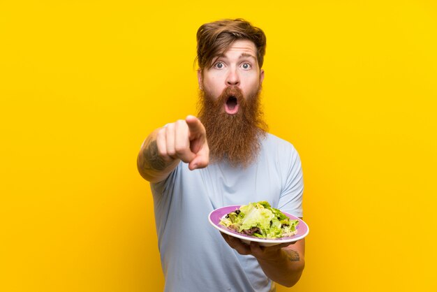 redhead man with long beard and with salad over isolated yellow wall surprised and pointing front