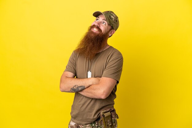 Redhead Military man with dog tag isolated on yellow background looking up while smiling