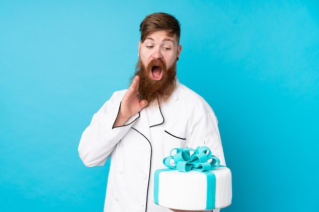 Redhead pastry chef with long beard holding a big cake over blue wall with surprise and shocked facial expression