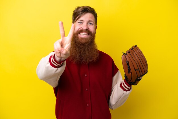 Redhead player man with beard with baseball glove isolated on yellow background smiling and showing victory sign