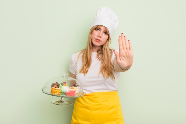 Redhead pretty woman looking serious showing open palm making stop gesture. home made cakes concept