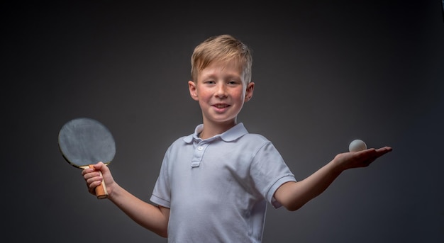 Redhead schoolboy dressed in a white t-shirt holds a ping-pong racquet and ball in a studio. Isolated on gray background.