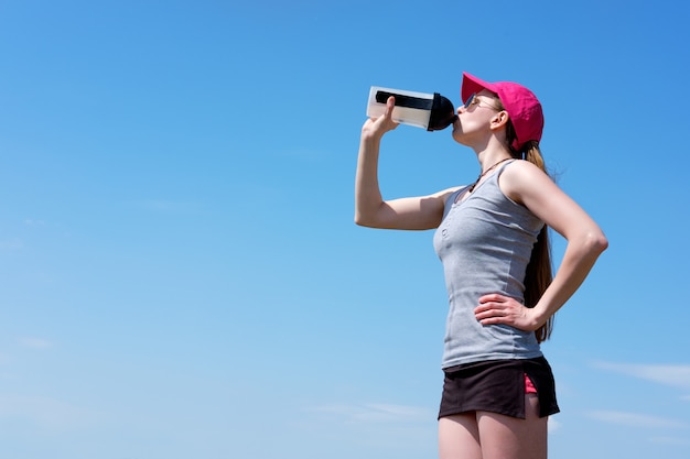 Redhead skinny girl in pink cap is drinking water after jogging, summer. Copy space
