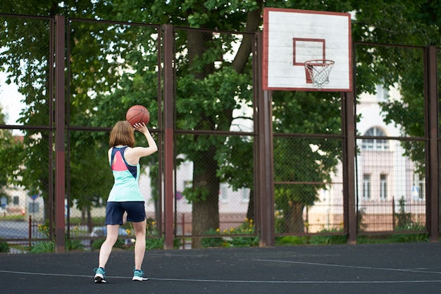 Рыжая худая девушка бросает мяч в баскетбольное кольцо на улице