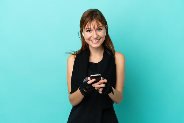 Redhead sport girl isolated on blue background sending a message with the mobile