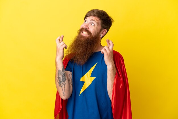 Redhead Super Hero man isolated on yellow background with fingers crossing
