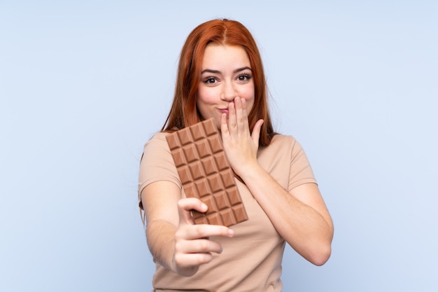 Redhead teenager girl over blue taking a chocolate tablet and surprised