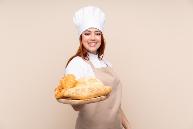 Redhead teenager girl in chef uniform. Female baker holding a table with several breads with happy expression