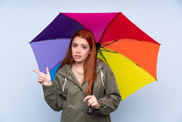Redhead teenager girl holding an umbrella over blue pointing to the laterals having doubts