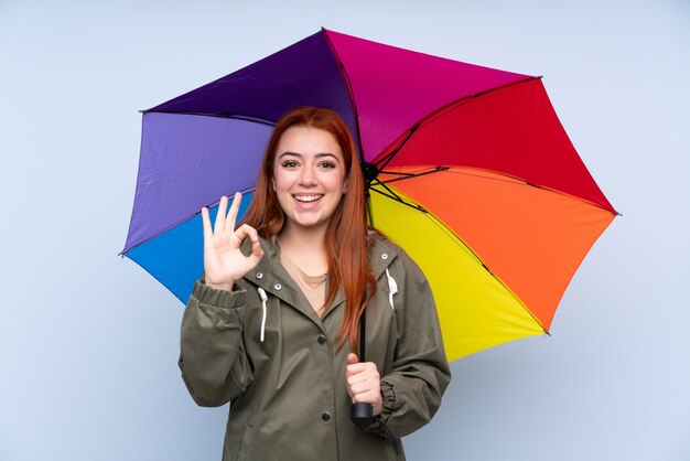 Redhead teenager girl holding an umbrella over blue showing an ok sign with fingers