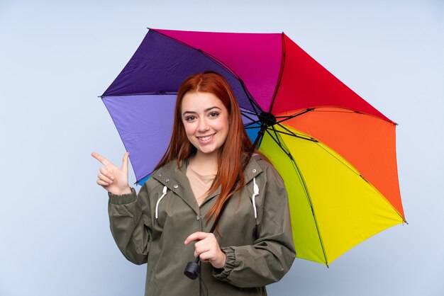 Redhead teenager girl holding an umbrella over isolated blue wall pointing finger to the side