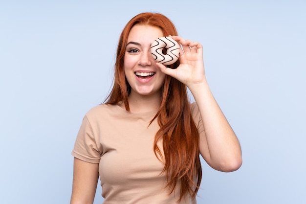 Redhead teenager girl over isolated blue wall holding a donut and happy