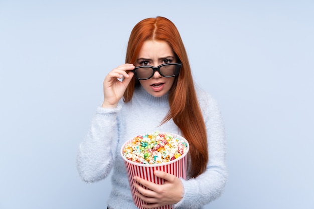 Redhead teenager girl over isolated blue wall surprised with 3d glasses and holding a big bucket of popcorns