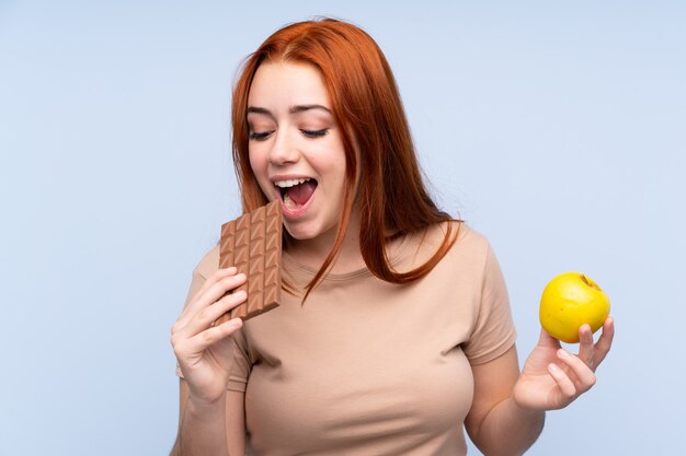 Redhead teenager girl over isolated blue wall taking a chocolate tablet in one hand and an apple in the other