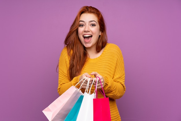 Photo redhead teenager girl over isolated purple wall holding shopping bags and surprised