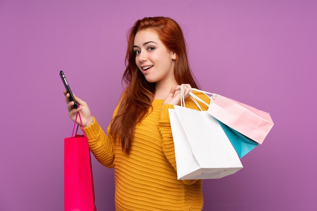 Redhead teenager girl over purple holding shopping bags and writing a message with her cell phone to a friend