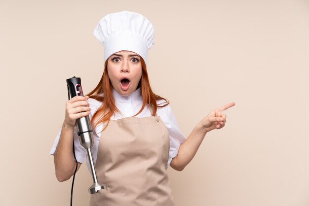 Redhead teenager girl using hand blender over isolated wall surprised and pointing side