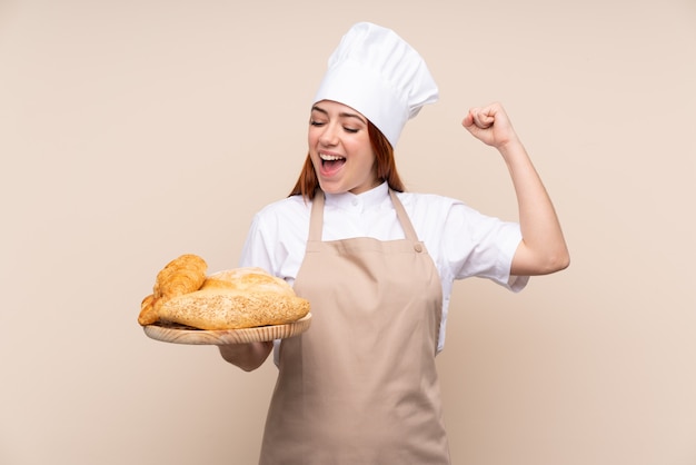 Redhead teenager woman in chef uniform. Female baker holding a table with several breads celebrating a victory