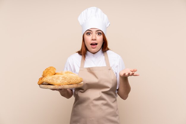 Redhead teenager woman in chef uniform. Female baker holding a table with several breads with shocked facial expression