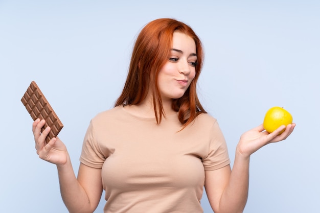 Redhead teenager woman having doubts while taking a chocolate tablet in one hand and an apple in the other