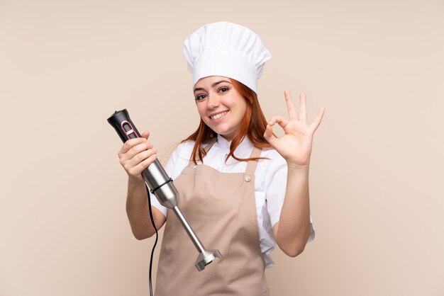 Redhead teenager woman using hand blender over isolated wall showing ok sign with fingers