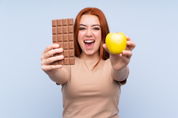 Redhead tienervrouw die een chocoladetablet in één hand en een appel in de andere nemen