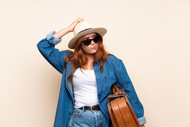 Redhead traveler girl with suitcase over isolated  having doubts and with confuse face expression