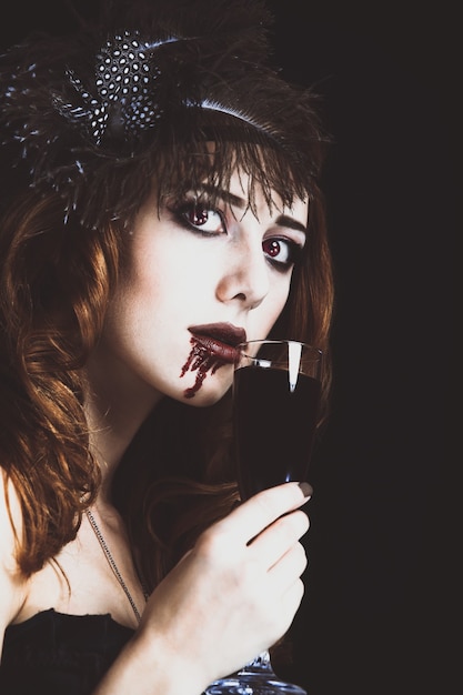 Redhead vampire woman with glass of blood. Photo in vintage style.