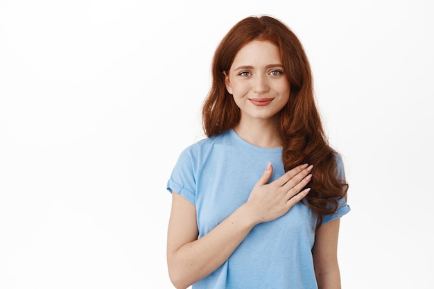 Redhead woman holding hand on heart and smiling, wearing blue t-shirt, standing over white background.