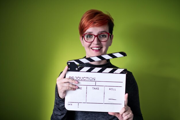 redhead woman holding movie clapper isolated against green background cinema concept in studio