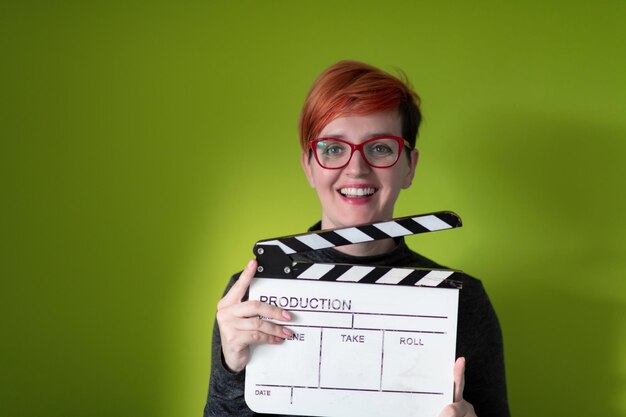 redhead woman holding movie clapper isolated against green background  cinema concept in studio