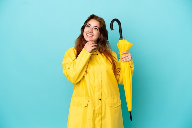 Redhead woman holding an umbrella isolated on blue background looking up while smiling