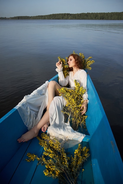 Redhead woman in white negligee dress sit in blue boat on pond lake. Red-haired girl with yellow flower branches sitting in boat at sunset