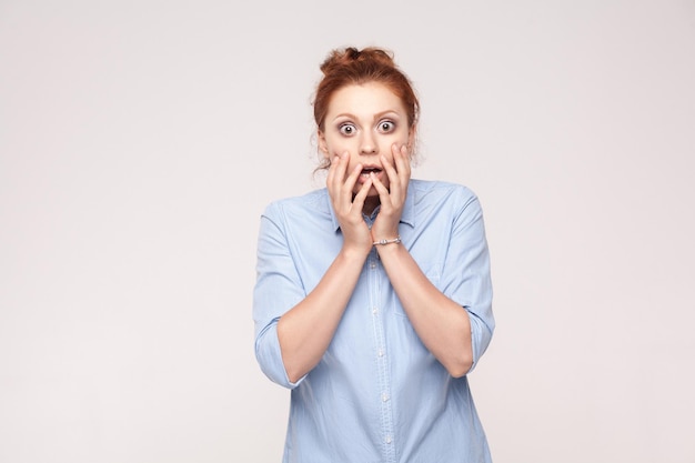 Redhead young adult woman holding arms on her cheeks and shocked