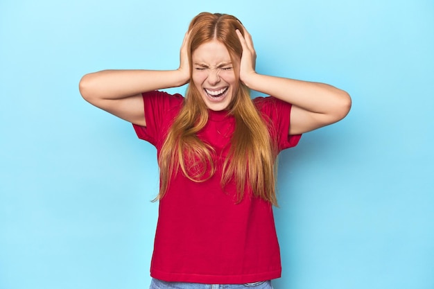 Redhead young woman on blue background covering ears with hands trying not to hear too loud sound