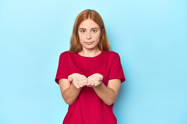 Redhead young woman on blue background holding something with palms offering to camera