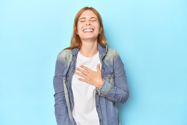 Redhead young woman on blue background laughs out loudly keeping hand on chest