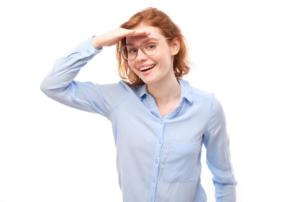 Redhead young woman in business shirt looks future looking far away distance with hand over head isolated on white background Search concept