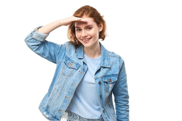 Redhead young woman in denim jacket looks future looking far away distance with hand over head isolated on white background Search concept