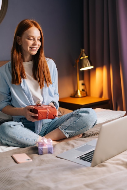 Redhead young woman holding gift box with present during online communication using laptop computer Happy lady sitting on bed at light bedroom and using laptop looking on display screen