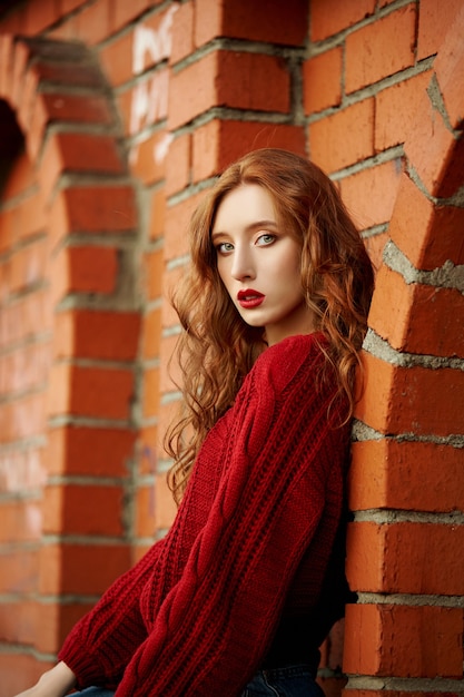 La giovane donna della testarossa in un maglione rosso cammina nel parco. ritratto di bellezza autunnale di una donna dai capelli rossi alla moda al tramonto