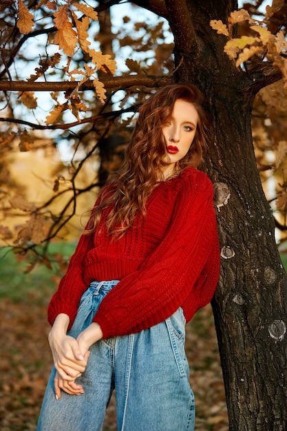 Redhead young woman in a red sweater walks in the park. Autumn beauty portrait of a fashionable Red-haired woman at sunset