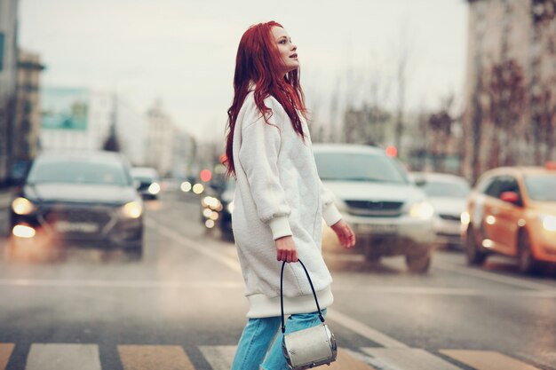 Redhead young woman in a white coat and blue jeans crosses the road at a pedestrian crossing.