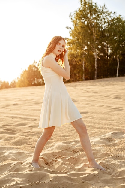 자연에서 여름에 모래에 맨발로 서 있는 흰 드레스에 빨간 머리 젊은 여자