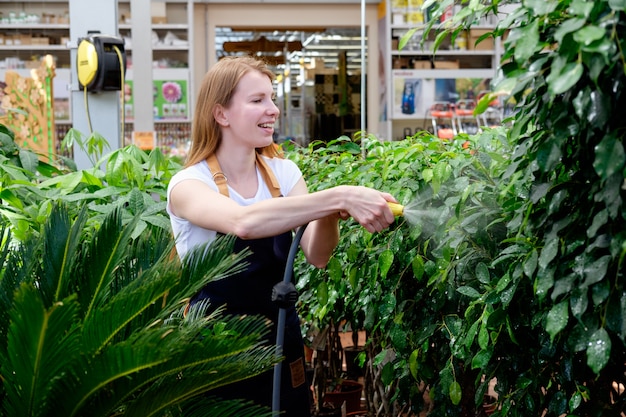 Redhead young woman worker in plant market greenhouse pouring plants and smiling