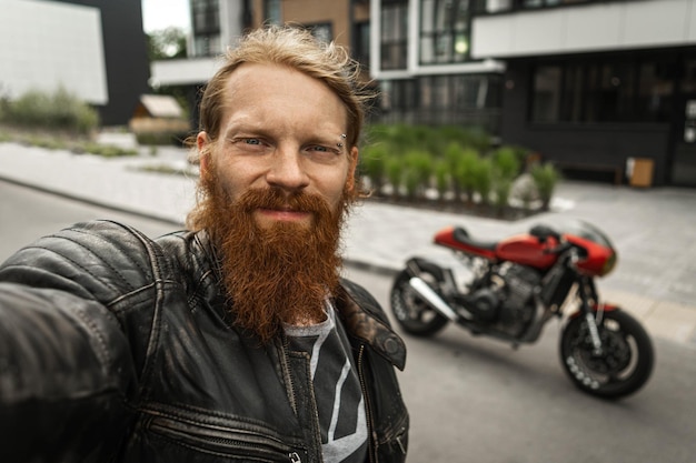 Foto motociclista barbuto dai capelli rossi senza casco che si fotografa davanti a una motocicletta