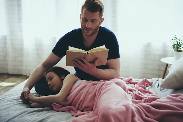 Redheaded single father reads book while daughter sleeps.