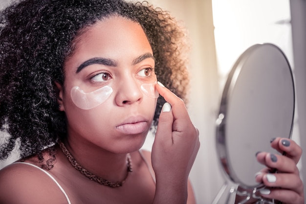 Reducing eye circles. Attentive dark-skinned woman checking effect of eye patches for decreasing eye bruises