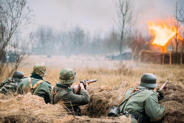 Reenactors armed rifles and dressed as world war ii german wehrmacht infantry soldiers fighting defensively in trench defensive position smokescreen building on fire on background