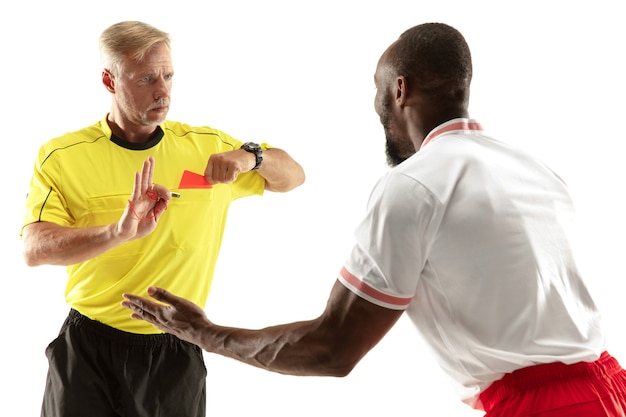 Referee showing a red card to a displeased african-american football or soccer player while gaming isolated on white wall. Concept of sport, rules violation, controversial issues, emotions.